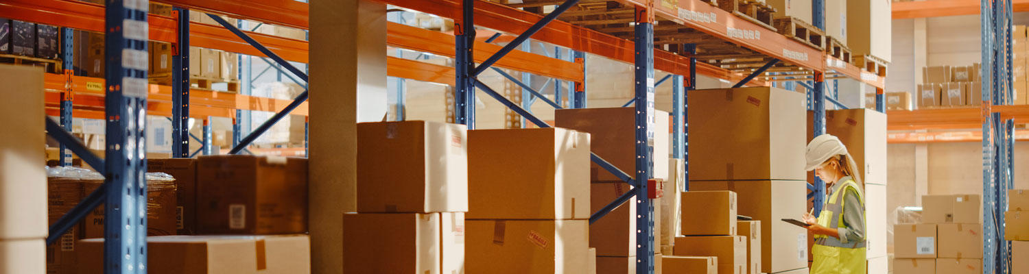 Photo showing a storage warehouse with tall shelves with lots of boxes and a woman employee checking off shipments of boxes.